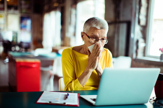 Senior woman blowing nose in tissue while working on office laptop
