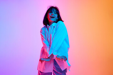 Dynamic photo of overjoyed young brunette woman dressed fashion outfit and dancing in neon light against gradient studio background. Concept of human emotions, fashion, beauty, style.