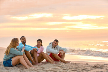 Young Couple With Friends In Casual Clothing On Vacation Sitting On Beach Watching Sunrise Together