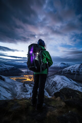 Man with backpack overlooking night city lights from peak - 774094153