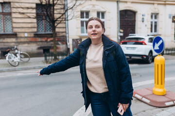 Stylish 30s woman with backpack holding smart phone and catching a taxi on the city street. Urban lifestyle concept.