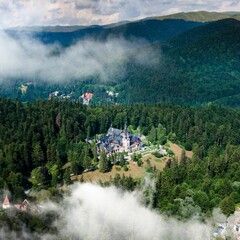 Aerial view of the Castle Peles surrounded by forested mountains in the clouds