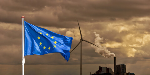Waving European flag in front of a polluting factory chimney with smoke and wind turbine - 774106747