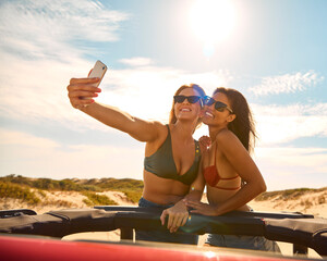 Two Women On Vacation In Car On Road Trip To Beach Standing Through Sun Roof Posing For Selfie