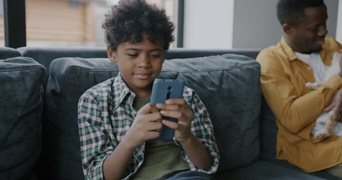 Cute African American boy using smartphone while father playing with cat sitting on sofa together. Modern gadget and family lifestyle concept.