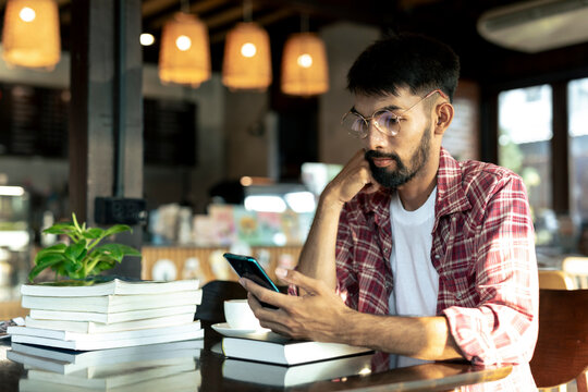 Portrait of young man with beard in glasses typing on his smart phone at cafe.