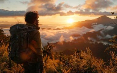A man with a backpack is standing on a mountain top, looking out at the sunset. The sky is filled with clouds, and the sun is setting in the distance