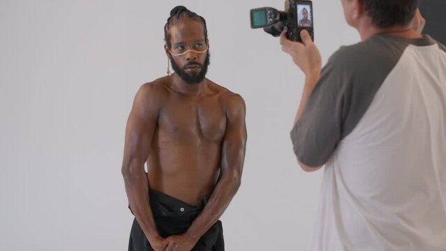 Muscular shirtless African American gay man with makeup poses for professional photographer in studio against white background. Photographer filming male model.