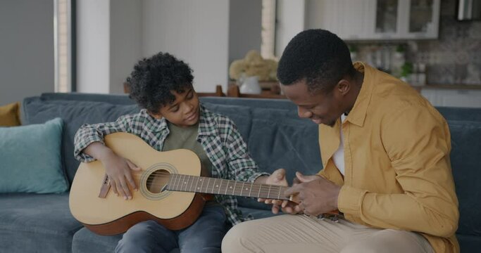African American man teaching child to play the guitar explaining bonding with son at home. Musical instrument and family relationship concept.