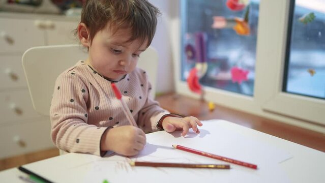 baby girl draws while sitting at a table by the window at home. happy family kid concept. baby daughter learns to draw with pencils on a sheet of dream paper indoors. development of fine motor skills