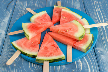 Slices of watermelon with popsicle sticks are laid out on a blue wooden background, creating a refreshing and colorful summer treat presentation