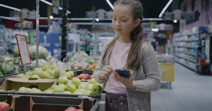 Portrait of adorable child using smartphone device shopping in supermarket buying fruit alone. Modern technology and retail concept.