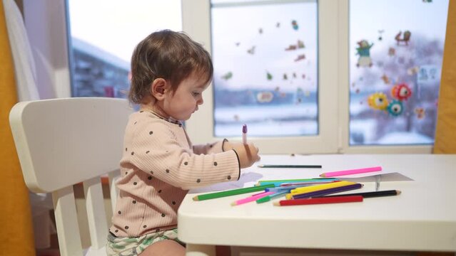 baby girl draws while sitting at a table by the window at home. happy family kid concept. dream baby daughter learns to draw with pencils on a sheet of paper indoors. development of fine motor skills