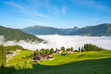 Morning clouds blanket the valley below a serene alpine landscape in Kitzbueheler Alps, with chalets and mountain peaks in view. Austria - 774150552