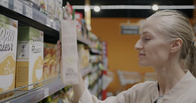 Cheerful young woman buying muesli in grocery store choosing organic food taking carton from shelf in supermarket. Consumerism and healthy nutrition concept.