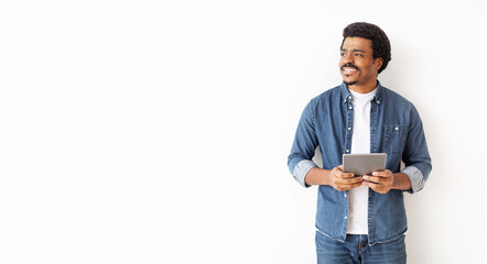 Relaxed black man in denim attire holding digital tablet and looking aside