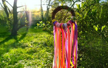 wreath with bright colorful ribbons in garden, green natural background. floral decor, Symbol of Beltane sabbat, Wiccan Celtic Holiday beginning of summer. pagan witch traditions, rituals - obrazy, fototapety, plakaty
