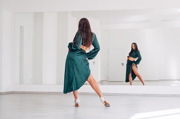 Graceful woman dressed in dark green Latin dancing dress doing elegant dance movings in white big hall with big mirror wall. People's expressions during dancing beauty of woman's body concept image