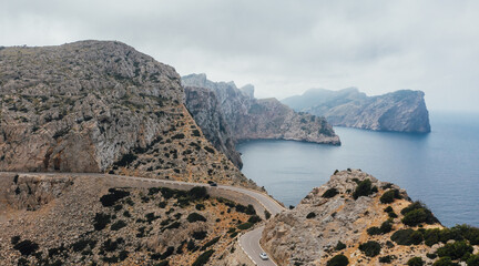 Lonely white Car on serpentine curved asphalt mountain road near Lighthouse of Cap de Formentor rocky coast. Mallorca Island, Balearic Islands, Spain. Flying drone aerial drone view shot.