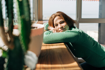A bored, relaxed red-haired freckled woman in a green long sleeve top in the rays of the summer sun by the window.