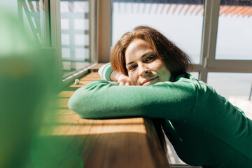 A bored, relaxed red-haired freckled woman in a green long sleeve top in the rays of the summer sun by the window.