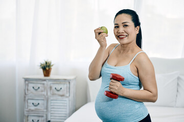 Smiling pregnant woman holding an apple and a dumbbell promoting healthy lifestyle in bedroom
