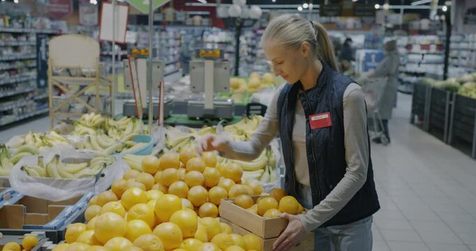 Female employee putting oranges on shelf working in supermarket preparing fruit for sale. Retail business and grocery store concept.