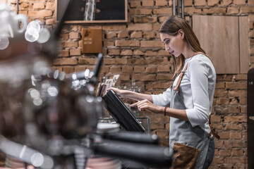Obraz na płótnie Canvas Young caucasian woman in apron working with cash box in coffee shop behind the counter. Female worker, barista, waitress, manager, administrator checking bill order on digital cash register.