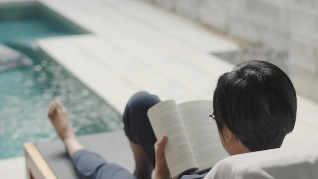 Back view and selective focus of beautiful asian adult woman who is reading a book beside the pool with tropical sunlight for refreshing with relaxation and comfort shows concept of summer vacation.