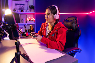 Host channel of beautiful Asian girl streamer with joystick playing online game wearing headphones pastel color talking with viewers media online. Esport skilled team players in neon room. Stratagem.