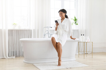 Woman in robe using phone by bathtub