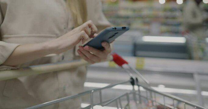 Close-up of female hands checking shopping list in smartphone application and touching food in trolley in supermarket. Modern gadget and consumerism concept.