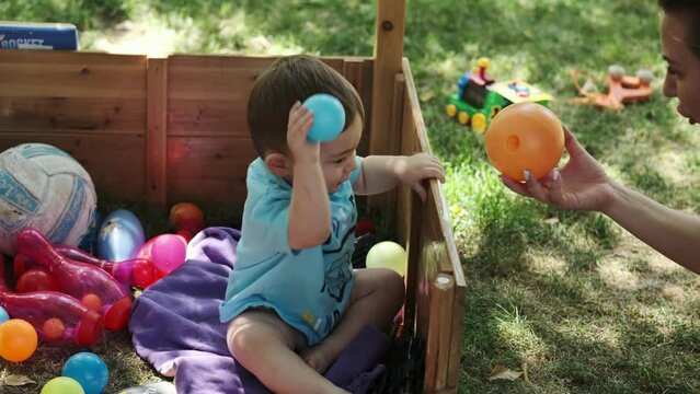 A little boy and his mother are playing with colorful balls in a wooden aviary on the lawn. Summer, sunny day