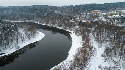 Drone photography of river flowing through public park and residential buildings near it covered by snow during winter day