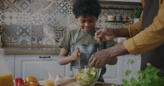 African American man cooking salad with adorable child preparing meal in kitchen at home. Family lifestyle and father son relationship concept.