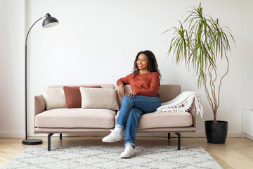 Relaxing Afternoon at Home: Smiling Black Woman on Sofa in Living Room - obrazy, fototapety, plakaty