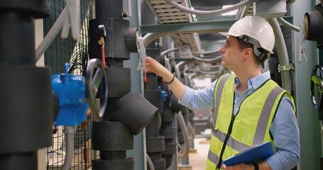 An industrial engineer in a hard hat and reflective vest inspecting machinery with clipboard paperwork in a manufacturing industrial facility.