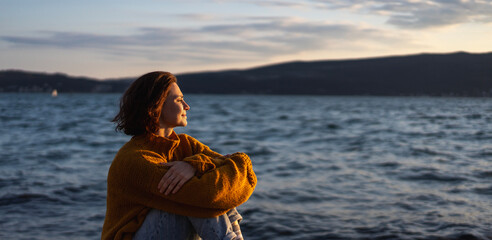 Smiling young woman in a yellow sweater looking at view at sunset enjoy sea view - 774246929