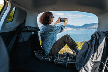 Young woman sitting in the open trunk of a car overlooking the sea with a smartphone in her hands, summer vacation and auto travel - 774247509