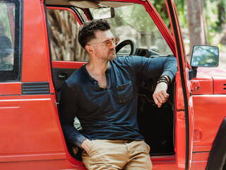 Attractive man and a red SUV against a background of palm trees. Sunny, clear day. Closeup, outdoor. Vacation and travel concept © Svetlana