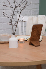 opened notebook with white pen, a cup of tea, burning candle, vase with branches on wooden table and chair. Top view.