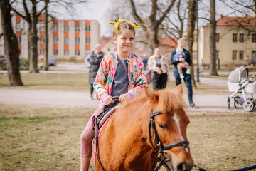 Valmiera, Latvia, April 1. 2024 - A smiling child on a pony holds the reins, wearing a colorful...