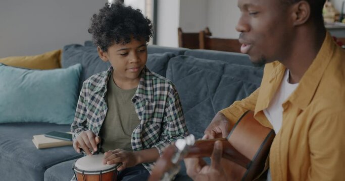Cute African American boy playing the drum while father adult man playing the guitar having fun at home. Family relationship and youth culture concept.