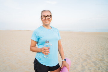 Cheerful sporty middle aged woman smiling at camera, holding water bottle and mat on the beach after workout training or active yoga on sunset. Female resting after fitness. Health and wellbeing.