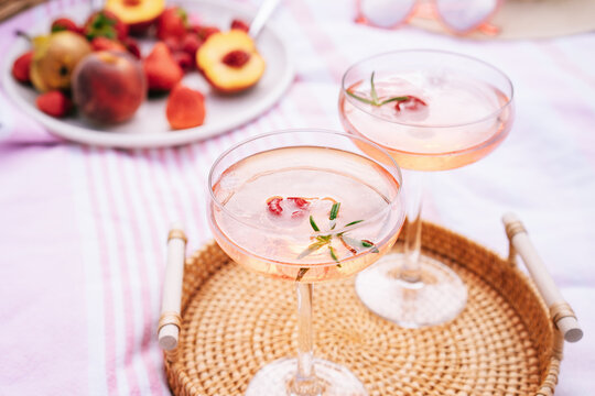 Pink drinks, cocktail with ice, raspberry, rosemary. Two glasses with martini, champagne, cider, lemonade on the blanket with fruit plate, picnic basket, Cozy summer picnic on nature. Selective focus.