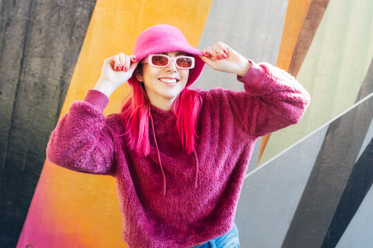 Hipster young woman with pink hair and sunglasses in magenta fluffy sweatshirt and bucket hat posing on the wall background. Urban street fashion. Mono color look. Gen Z, millennials self expression.