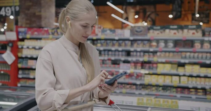 Young lady shopping in supermarket using smartphone app and looking at food in trolley. Modern technology and consumerism concept.
