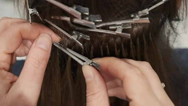 Fusion hair extensions in a beauty salon using capsules and heating tool. Hairdresser installing strands of hair to brunette woman. Hair extension with keratin, increase hair volume