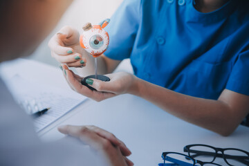 Close-up of Asian female doctor talking with elderly patient showing eyeball model and explaining...