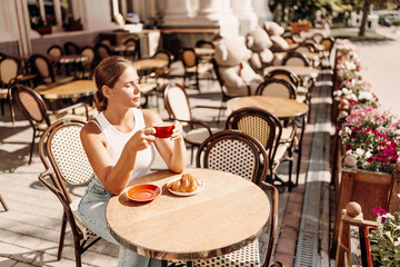 Portrait of happy woman sitting in a cafe outdoor drinking coffee. Woman while relaxing in cafe at table on street, dressed in a white T-shirt and jeans